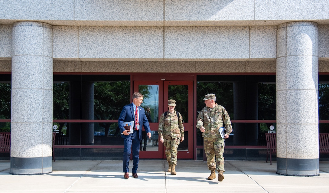 Three people exit a large stone building. Two are in camoflauge uniforms and one is in a blue suit. Two men and a woman.