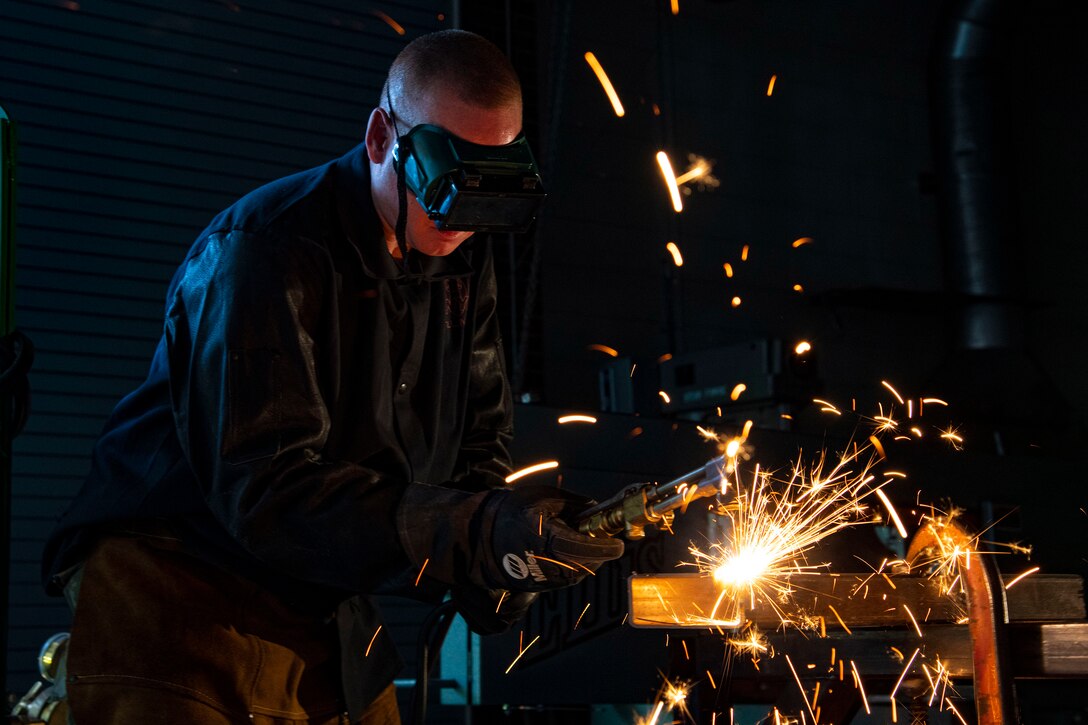 A service member wearing goggles uses a device to cut through metal, creating sparks.