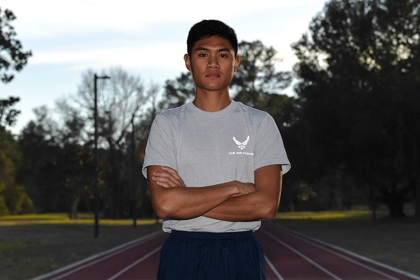 A stoic-looking man stands with his arms crossed over his chest while standing on a running track.