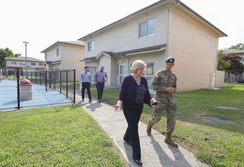 A group of four people, including one uniformed service member, walk down a sidewalk in a neighborhood.