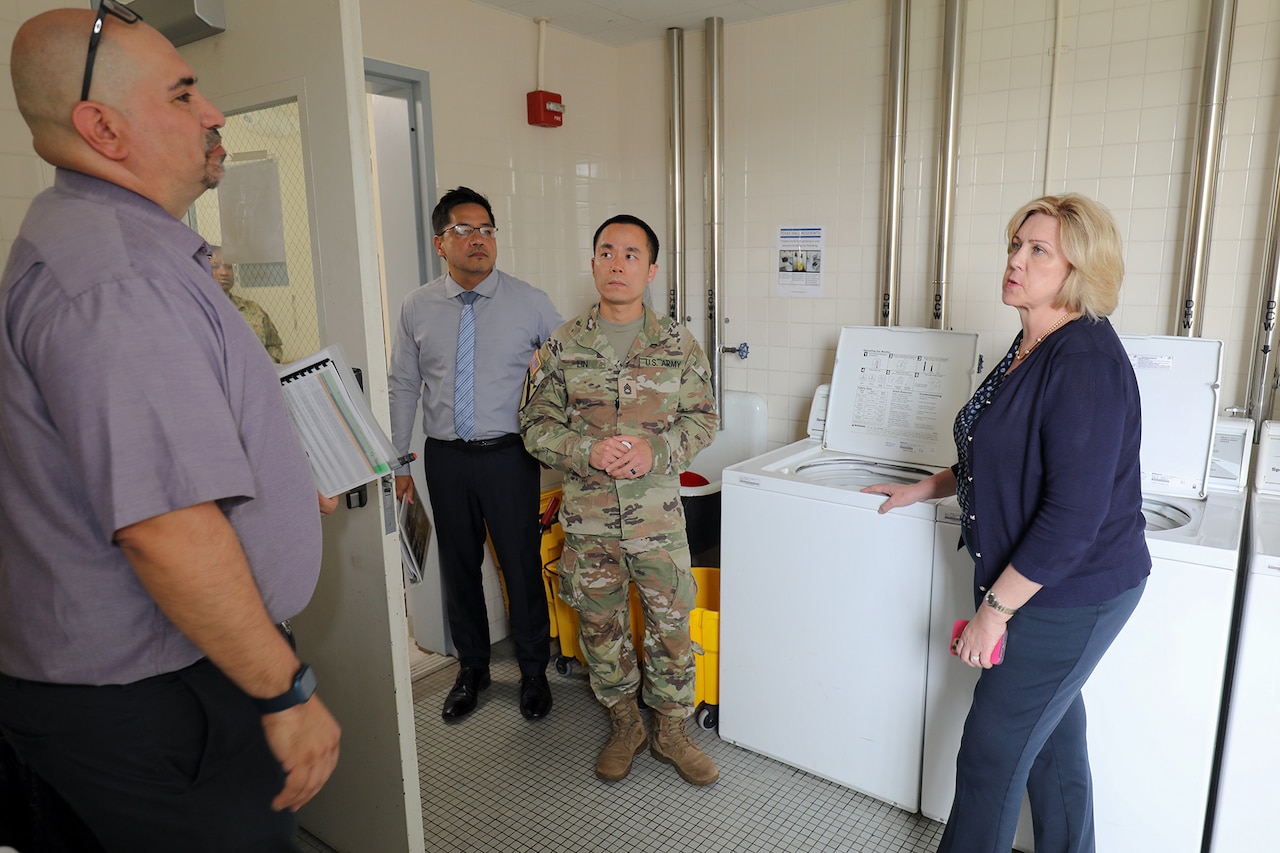A group of four people, including one uniformed service member, stand inside a laundry room.