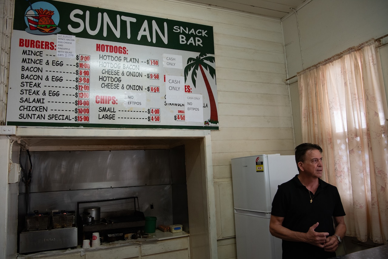 A man stands next to a grill under a menu displayed on a wall.