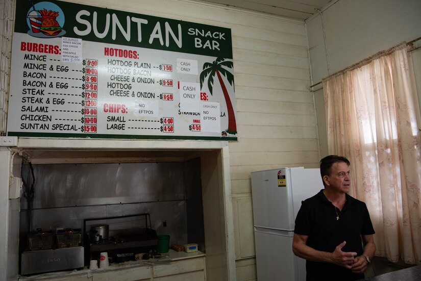 A man stands next to a grill under a menu displayed on a wall.