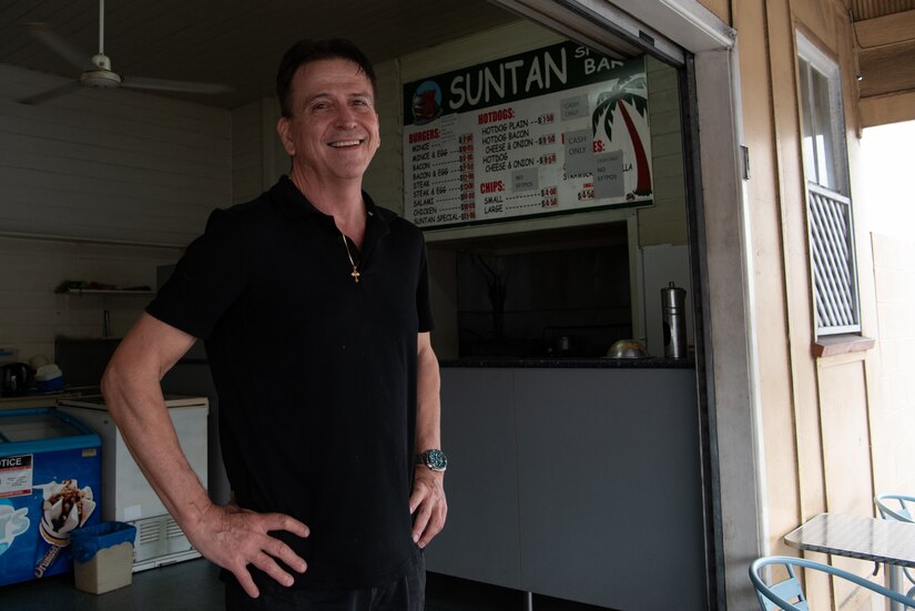 A man is photographed outside of a storefront with a menu displayed on a wall in the background.