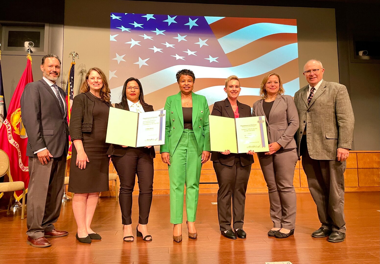 A group of people stand on a stage with an image of the American flag behind them.