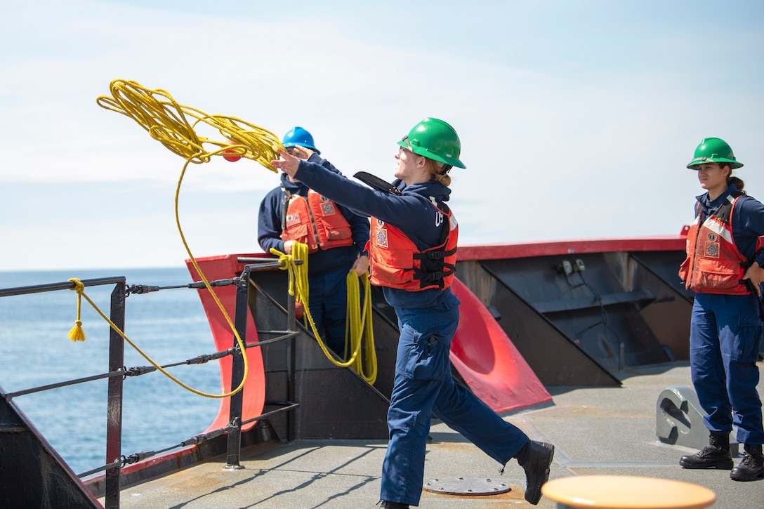 A Coast Guardsman throws a yellow line off of a ship as two fellow Coast Guardsmen watch.