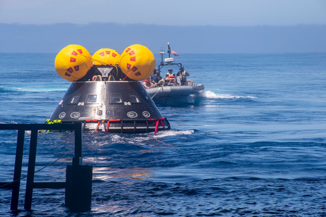 Sailors riding in a small boat displaying an American flag tow a space capsule in a body of water.