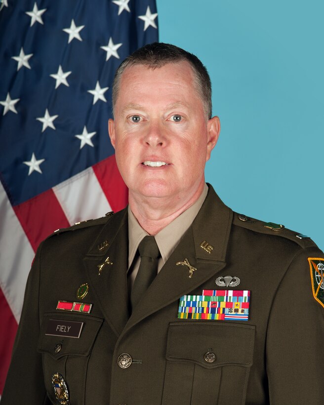 Official photo of Chief Warrant Officer 5 Jeffery Fiely in front of the U.S. flag and teal background.