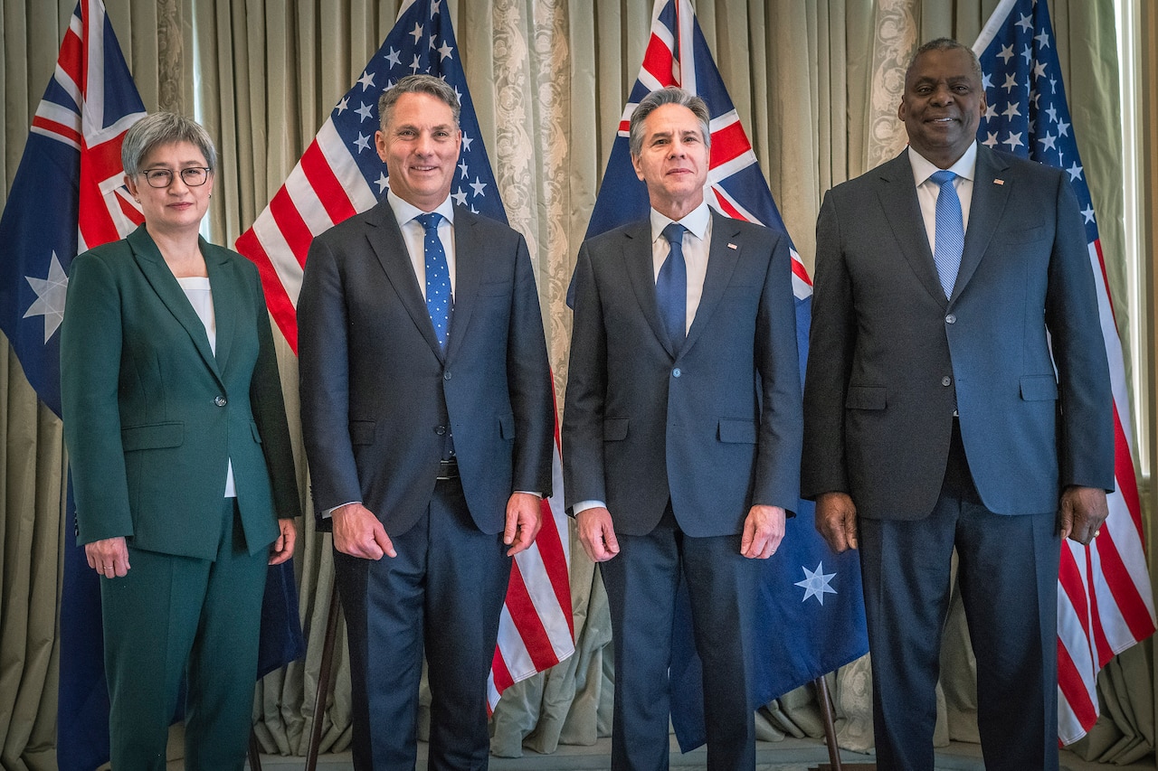 Four people in business attire smile for photographs while standing in front of flags.