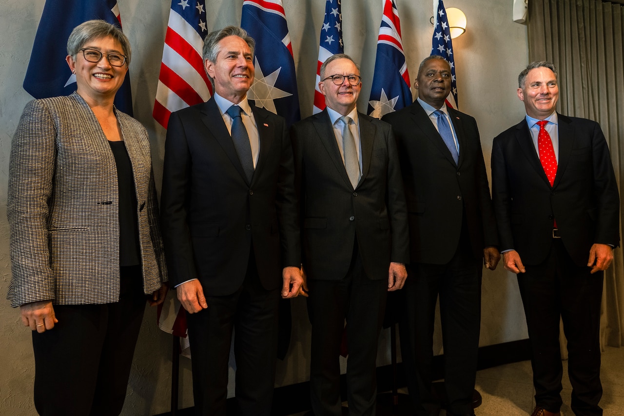 Five people in business attire smile and pose for a photo, standing in front of flags.