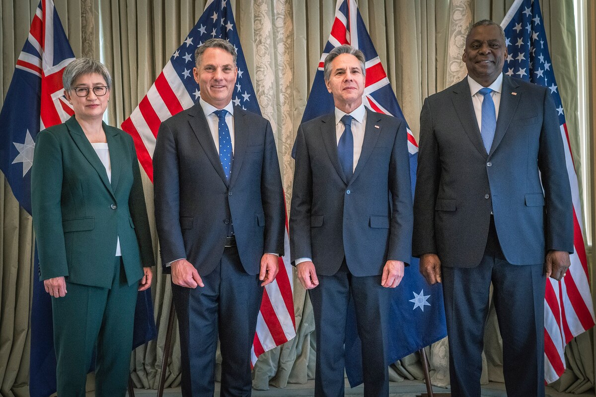 Four people in business attire smile for photographs while standing in front of flags.