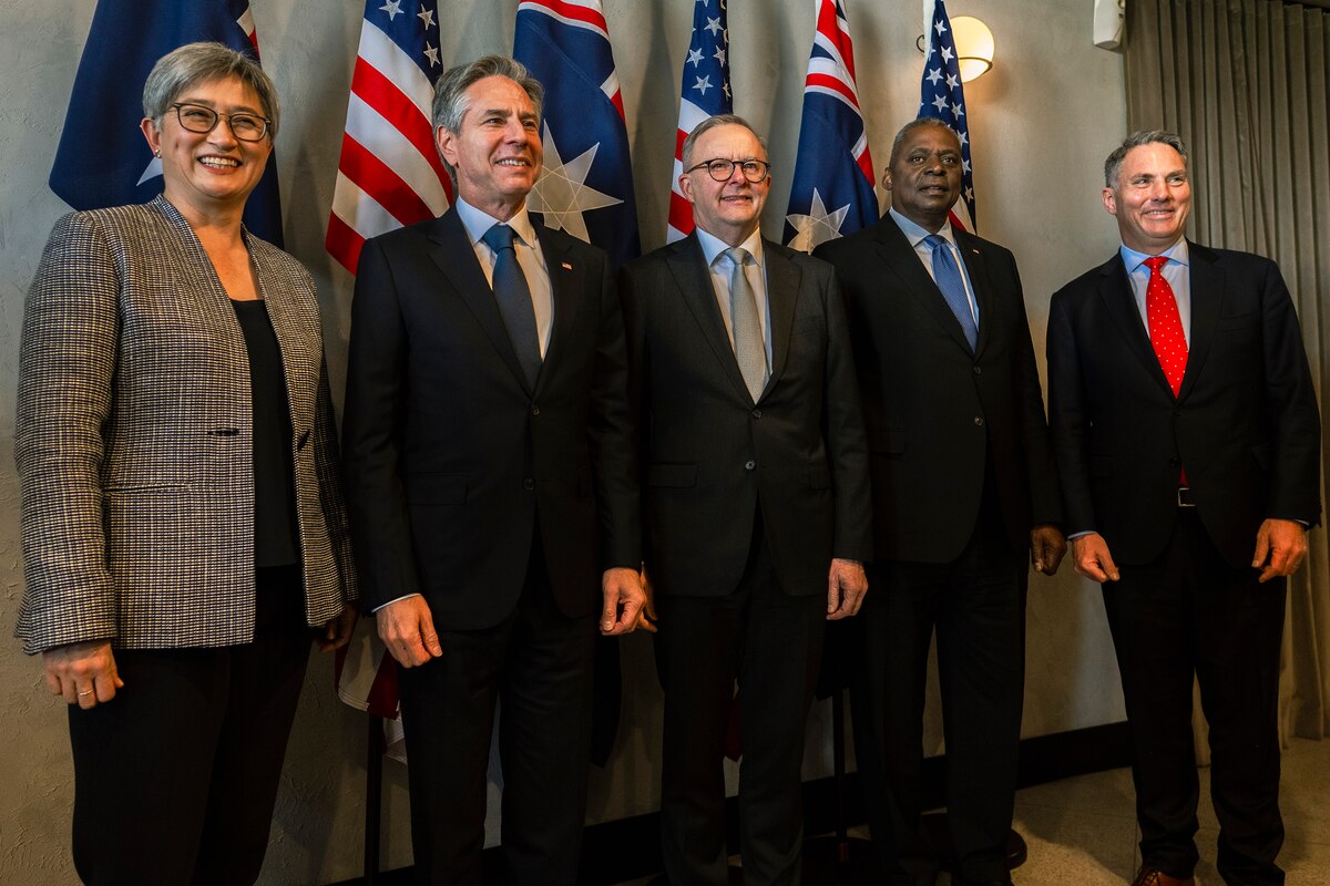 Five people in business attire smile and pose for a photo, standing in front of flags.