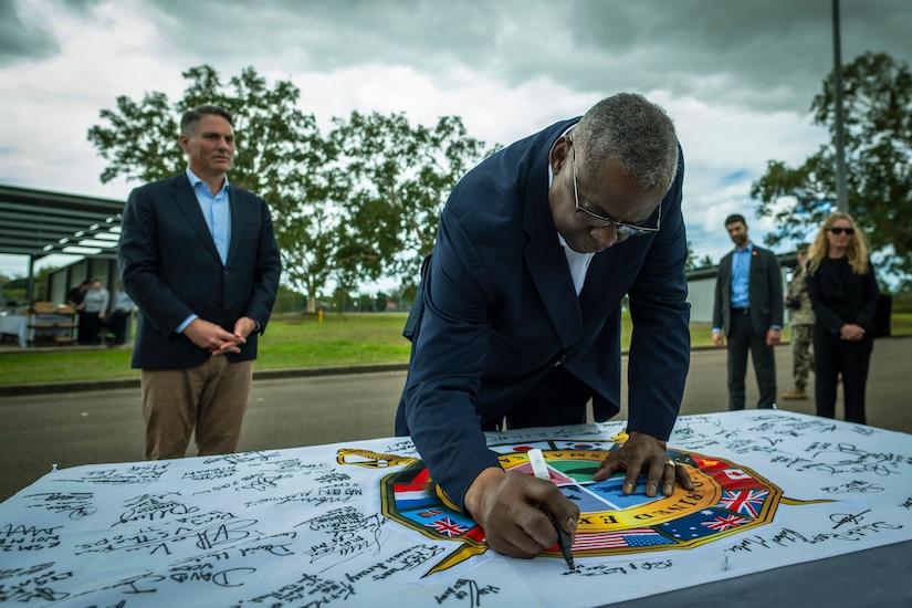 A person wearing a business suit leans over a table to sign a large flag containing many other signatures as another man looks on.