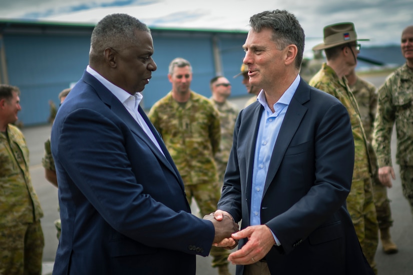 Two people wearing business suits shake hands, as service members in military camouflage uniforms mill about in the background.