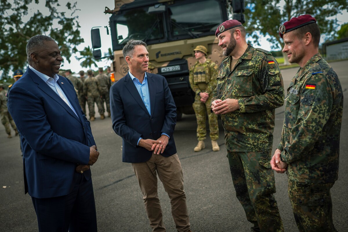 Two people wearing business suits stand outside and talk to two uniformed service members.