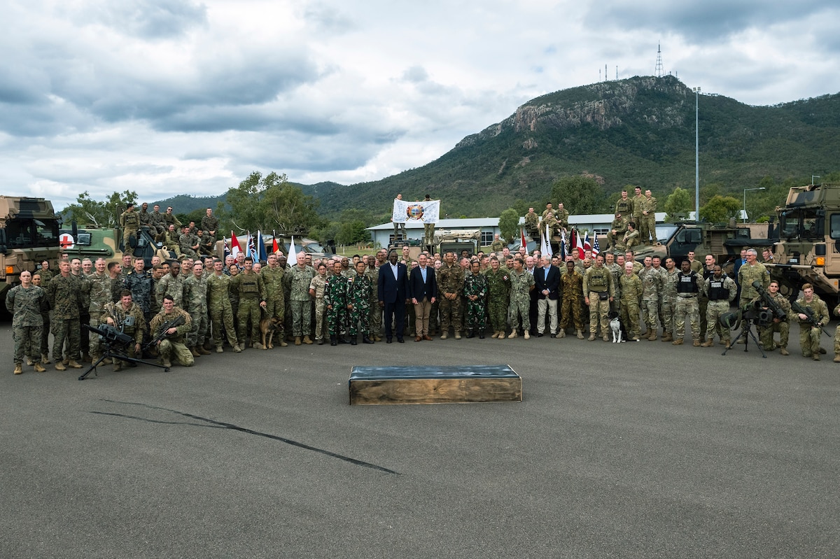 A large group of service members wearing camouflage uniforms pose for a photo. Three people wearing business suits pose with the troops.