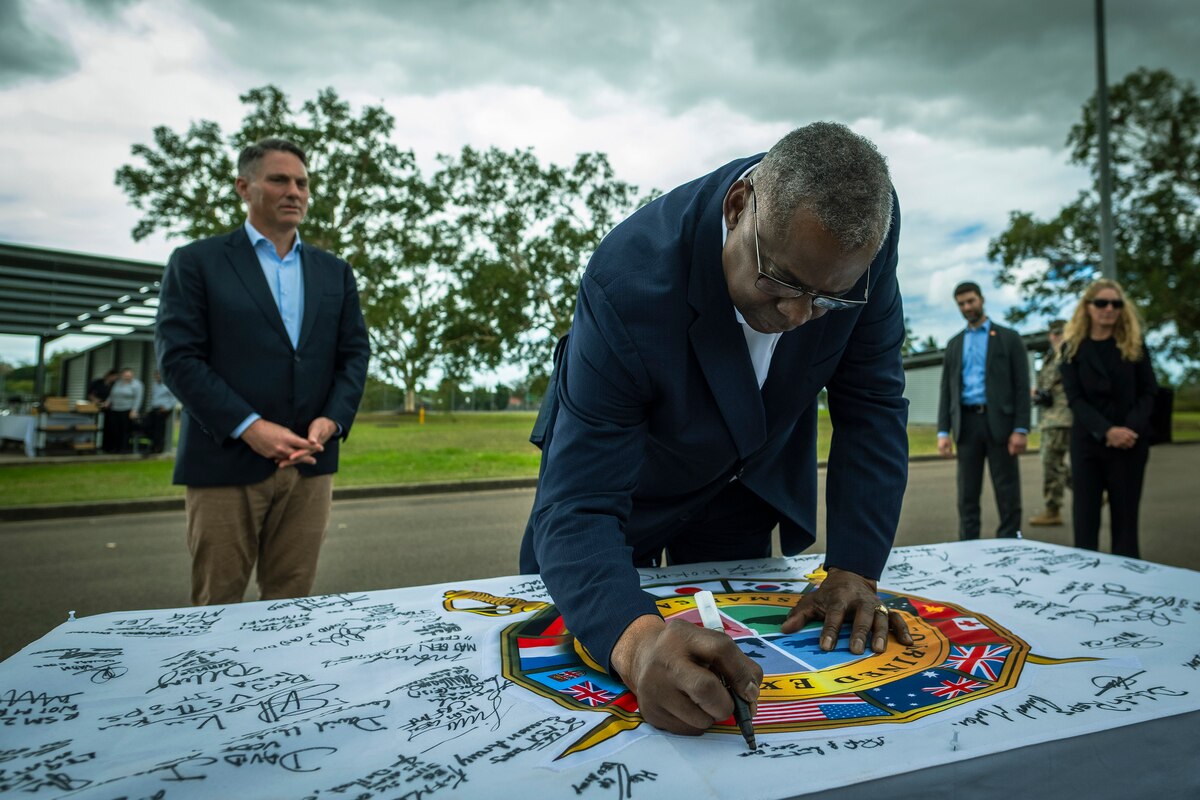 A person wearing a business suit leans over a table to sign a large flag containing many other signatures as another man looks on.