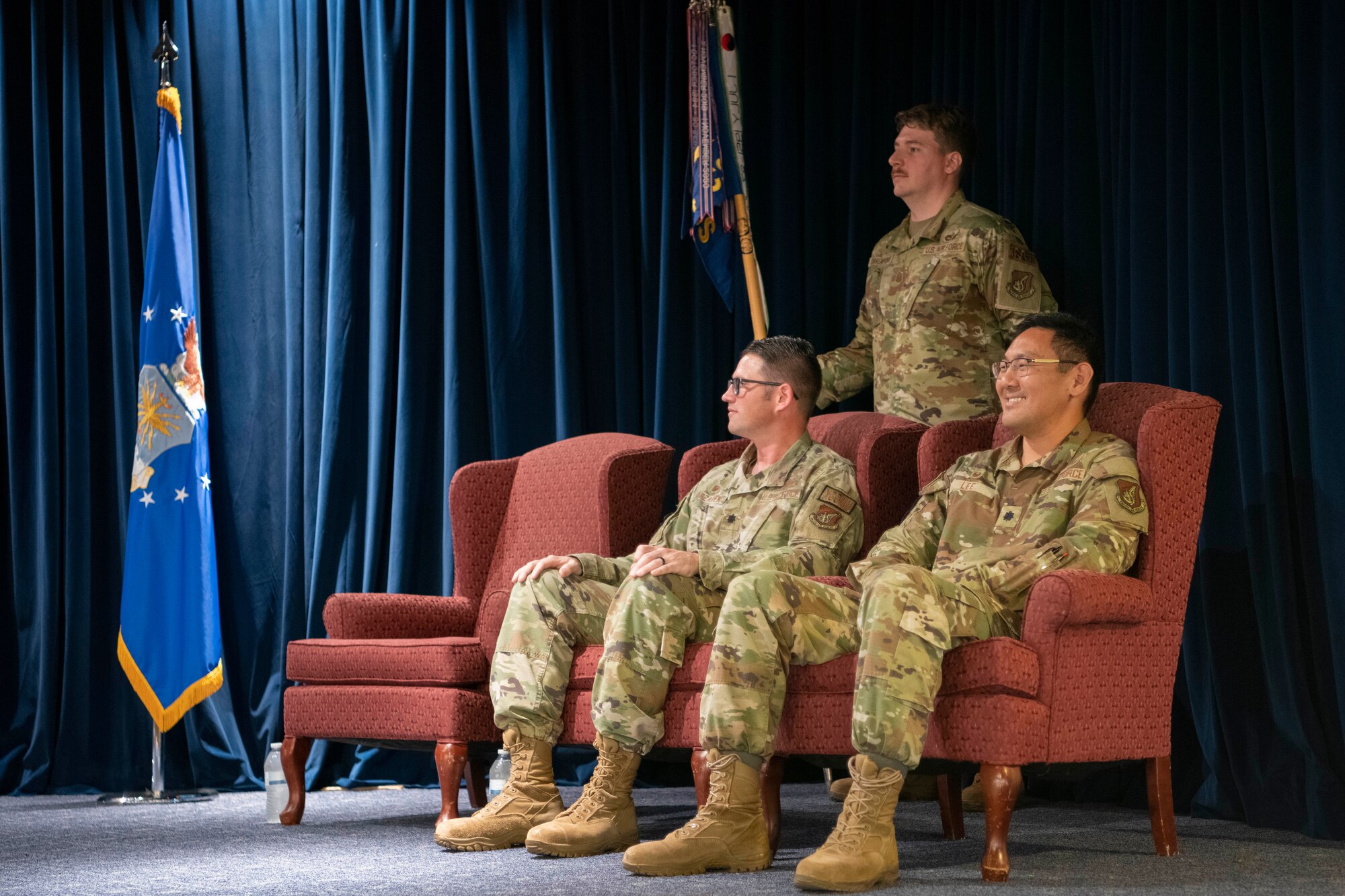 Two military men sit in chairs on stage.