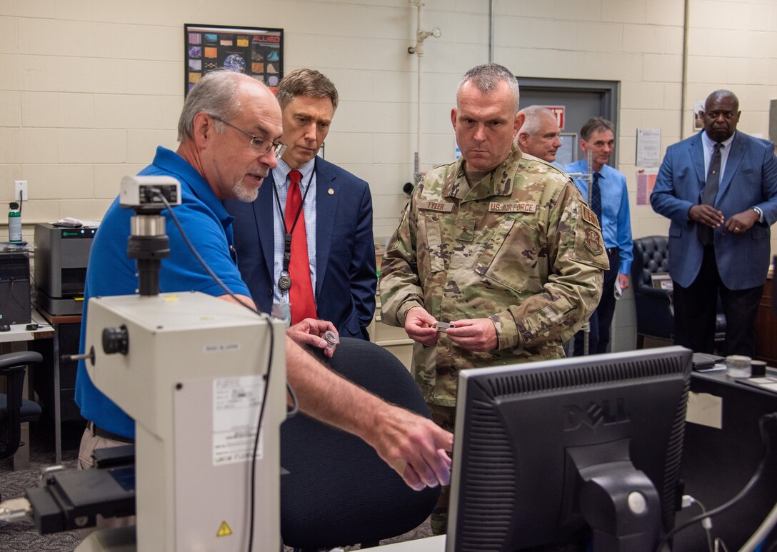 A man in military uniform stands next to a man in a blue suit watch another man in a blue shirt and glasses explain a procedure on a testing machine in a mechanical lab.