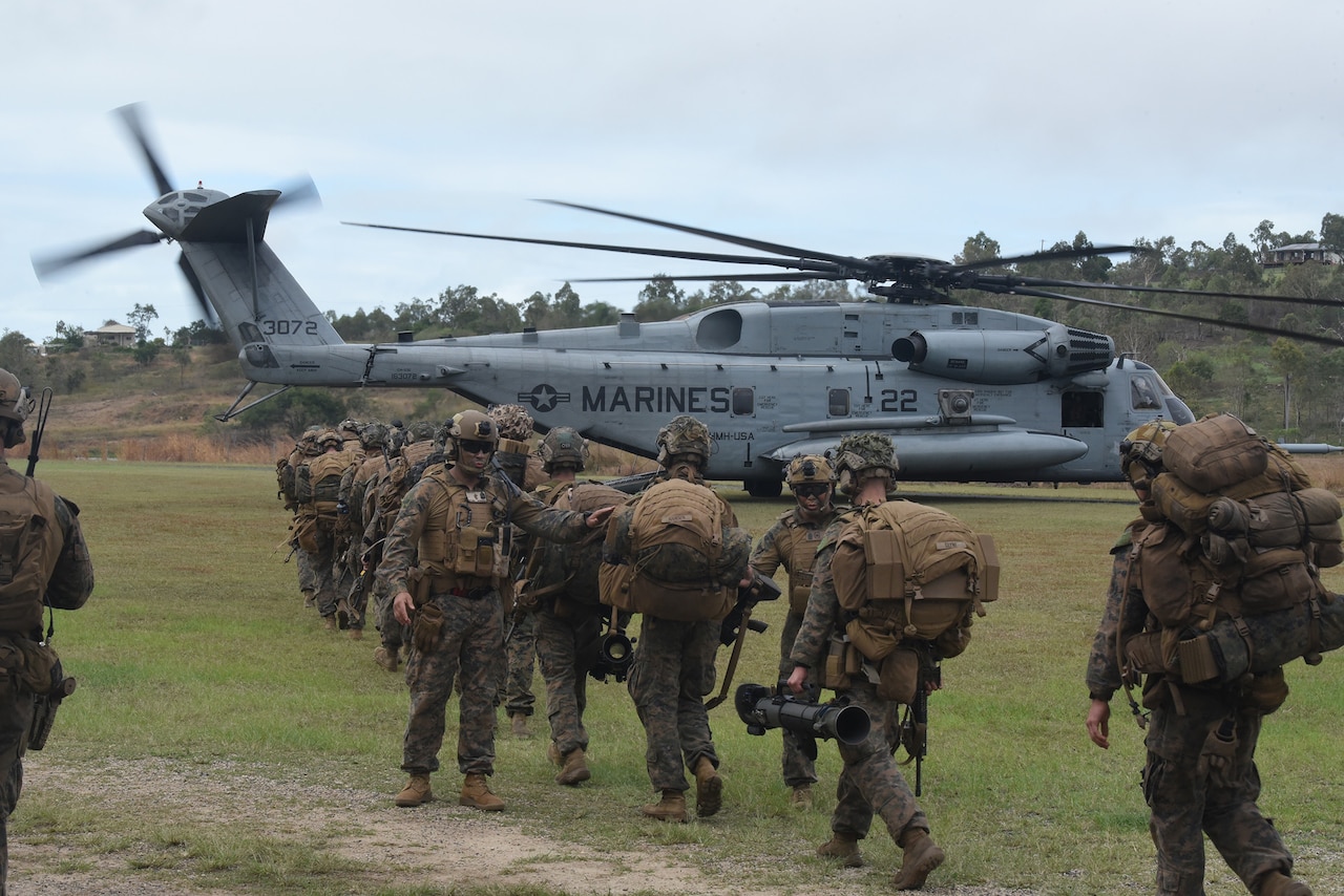 Marines dressed in camouflage uniforms and carrying heavy packs and gear walk to a helicopter.