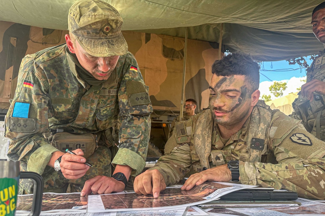 Two soldiers in battle gear point to maps laying on a table inside a tent.