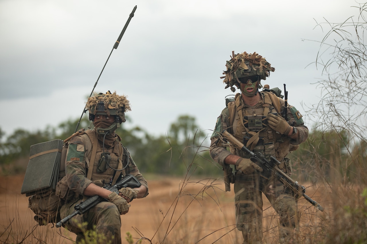 A soldier in battle gear kneels next to a fellow soldier while in the field.