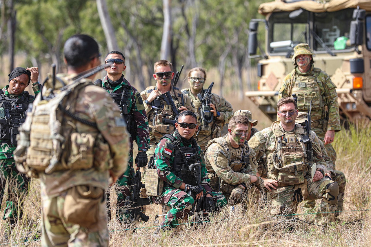 A group of soldiers in battle gear gathers around another soldier in a field.