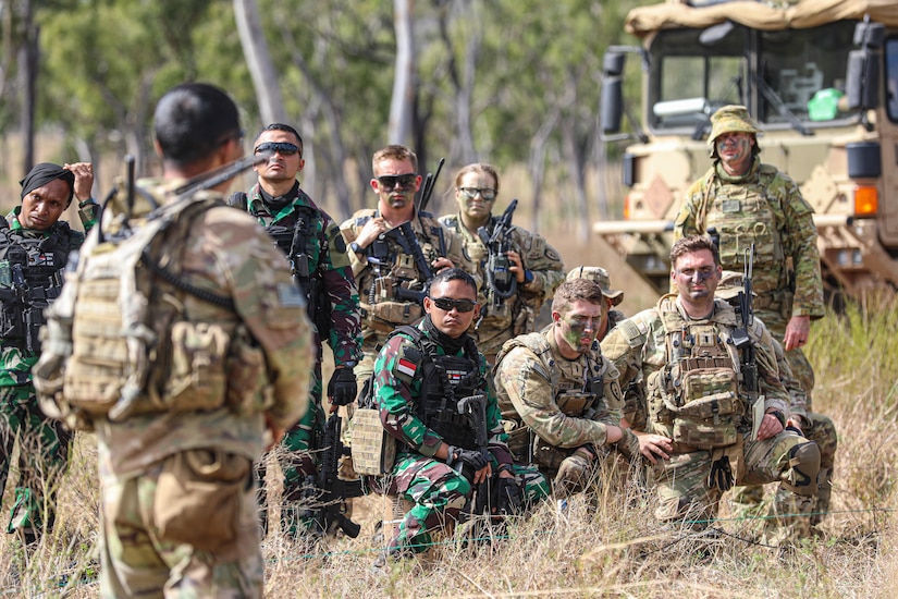 A group of soldiers in battle gear gathers around another soldier in a field.