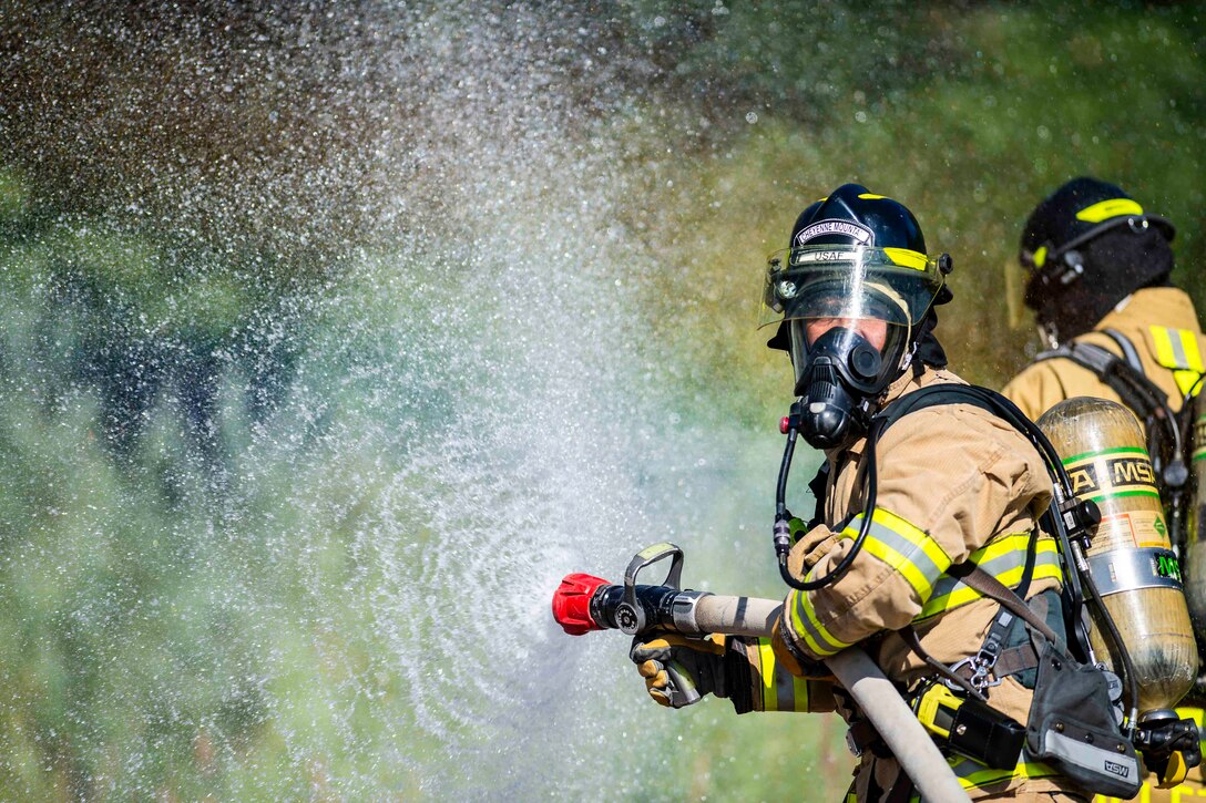 A person wearing fire protective equipment sprays water from a hose as another person stands behind.