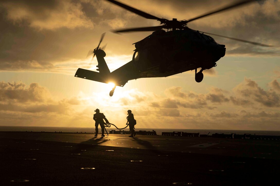 Two Marines shown in silhouette carry a rope on the deck of a ship as a helicopter flies above.