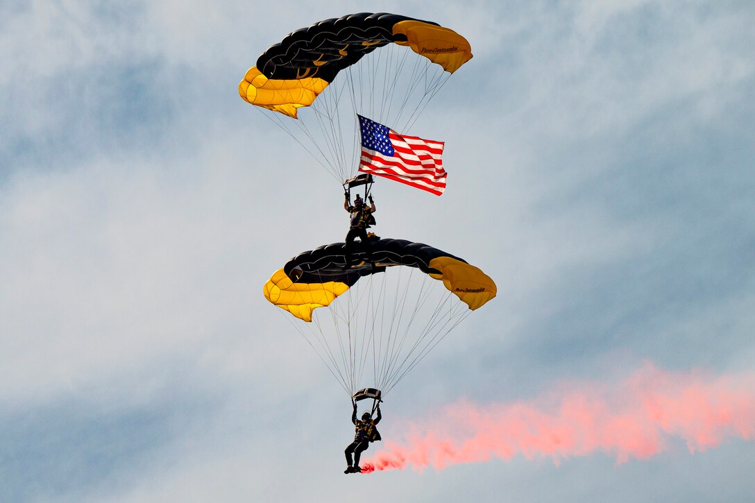One service member with a parachute stands on another’s parachute as they descend while displaying an American flag and red smoke.