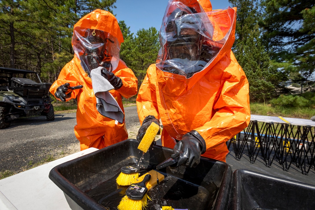 Soldiers wearing hazmat gear clean a camera with a sponge and rag in a black bin with greenery in the background.