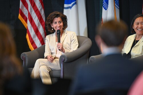 the secretary sits in a chair at the front of the room, holding a microphone. She smiles as an audience member engages with her off camera.