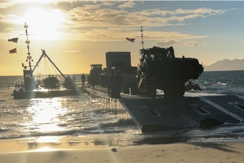 A large boat carrying heavy equipment makes landfall on a beach.