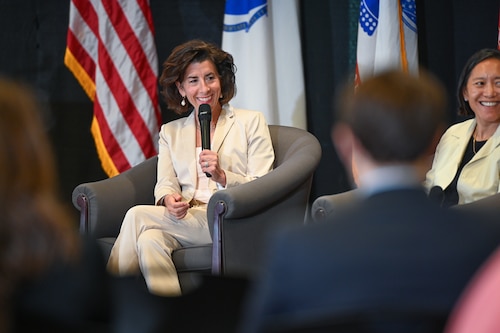the secretary sits in a chair at the front of the room, holding a microphone. She smiles as an audience member engages with her off camera.