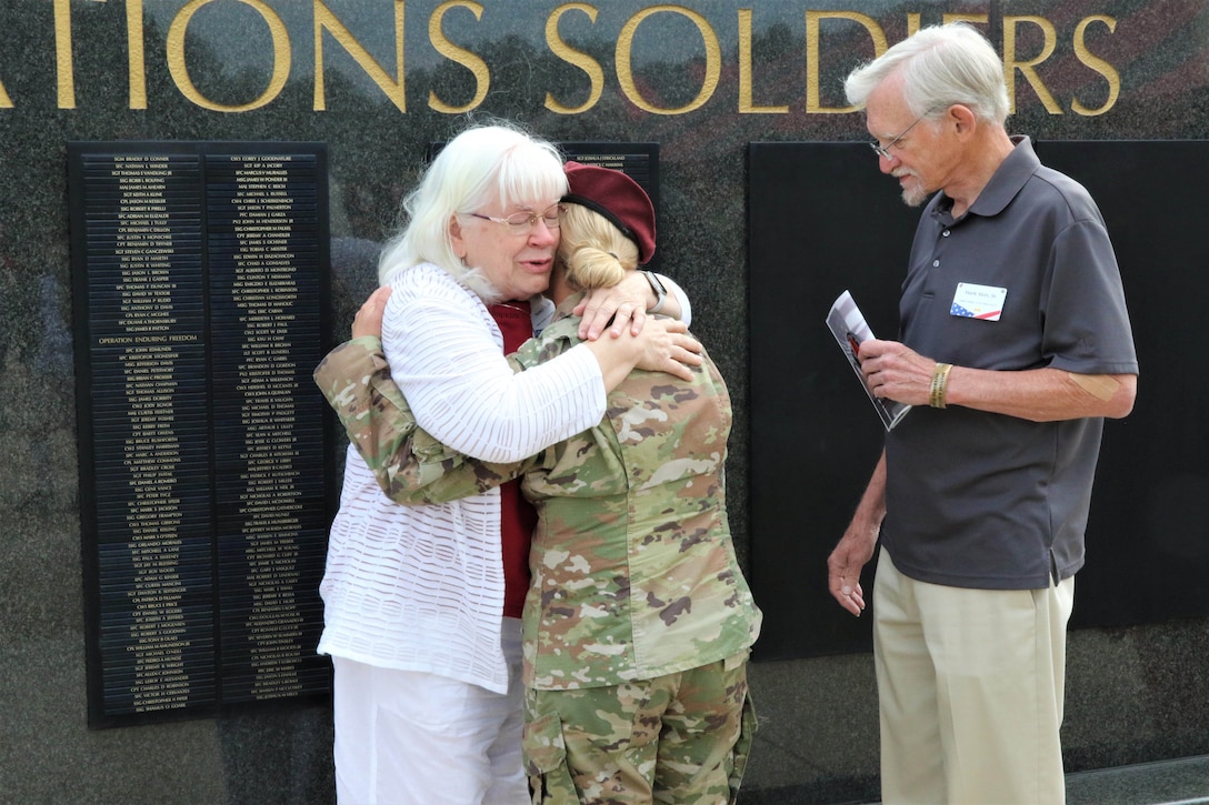 A soldier hugs a woman while a man stands next to them in front of a large wall.