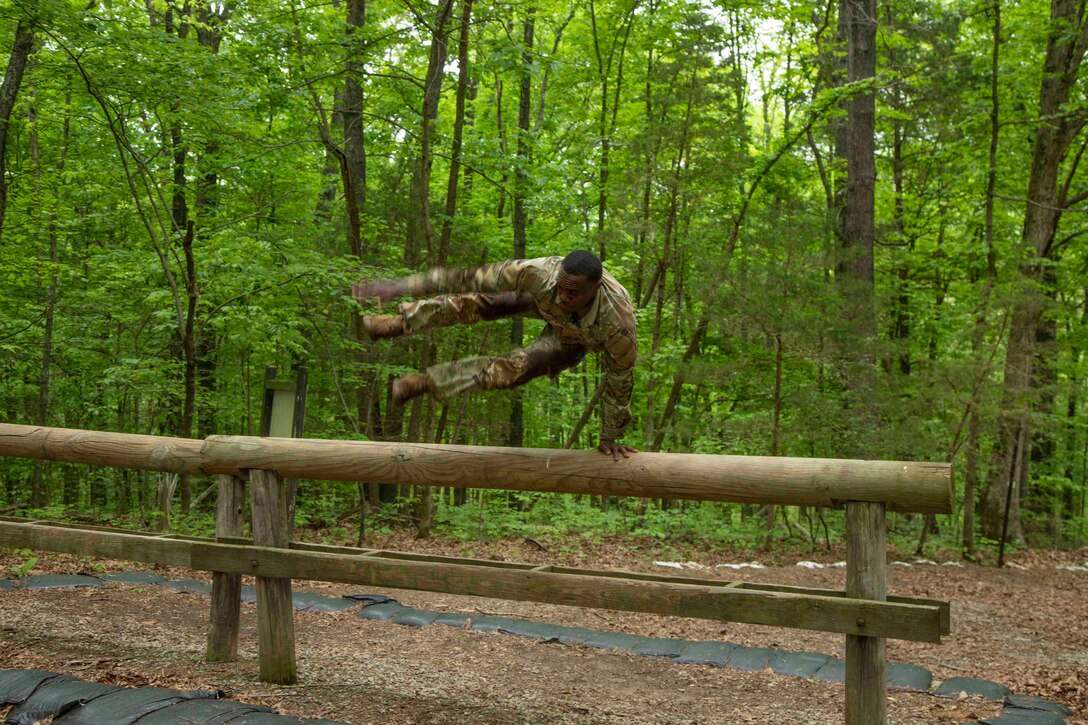 A soldier hops over a wooden obstacle in a wooded area.