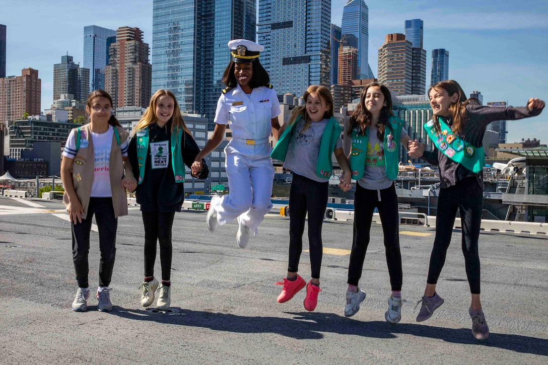 A sailor jumps in the air while holding hands with children; some of the children are also jumping.