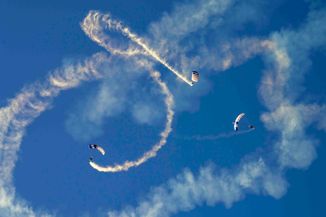 Three airmen descend in the sky wearing parachutes as white smoke trails from their feet.