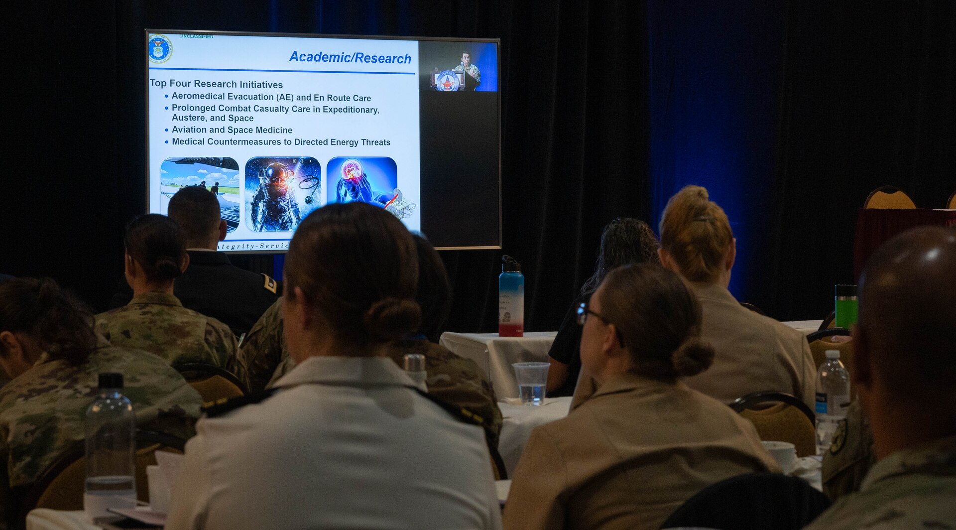 The TriService Nursing Research Program's annual Dissemination Course was held on April 4-6th, 2023, San Antonio, Texas. A room full of people sit while Brig. Gen. Jeannine Ryder, Chief Nurse of the Air Force, presents a  power point discussing the top four research initiatives: aeromedical evacuation (AE) and en route care, prolonged combat casualty care in expeditionary and space, aviation and space medicine, and medical countermeasures to directed energy threats.