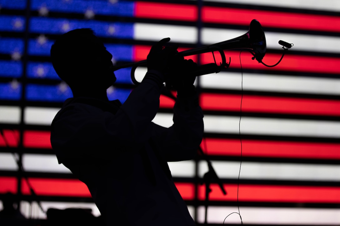 A sailor plays a trumpet in silhouette with a red, white and blue U.S. flag light in the background.