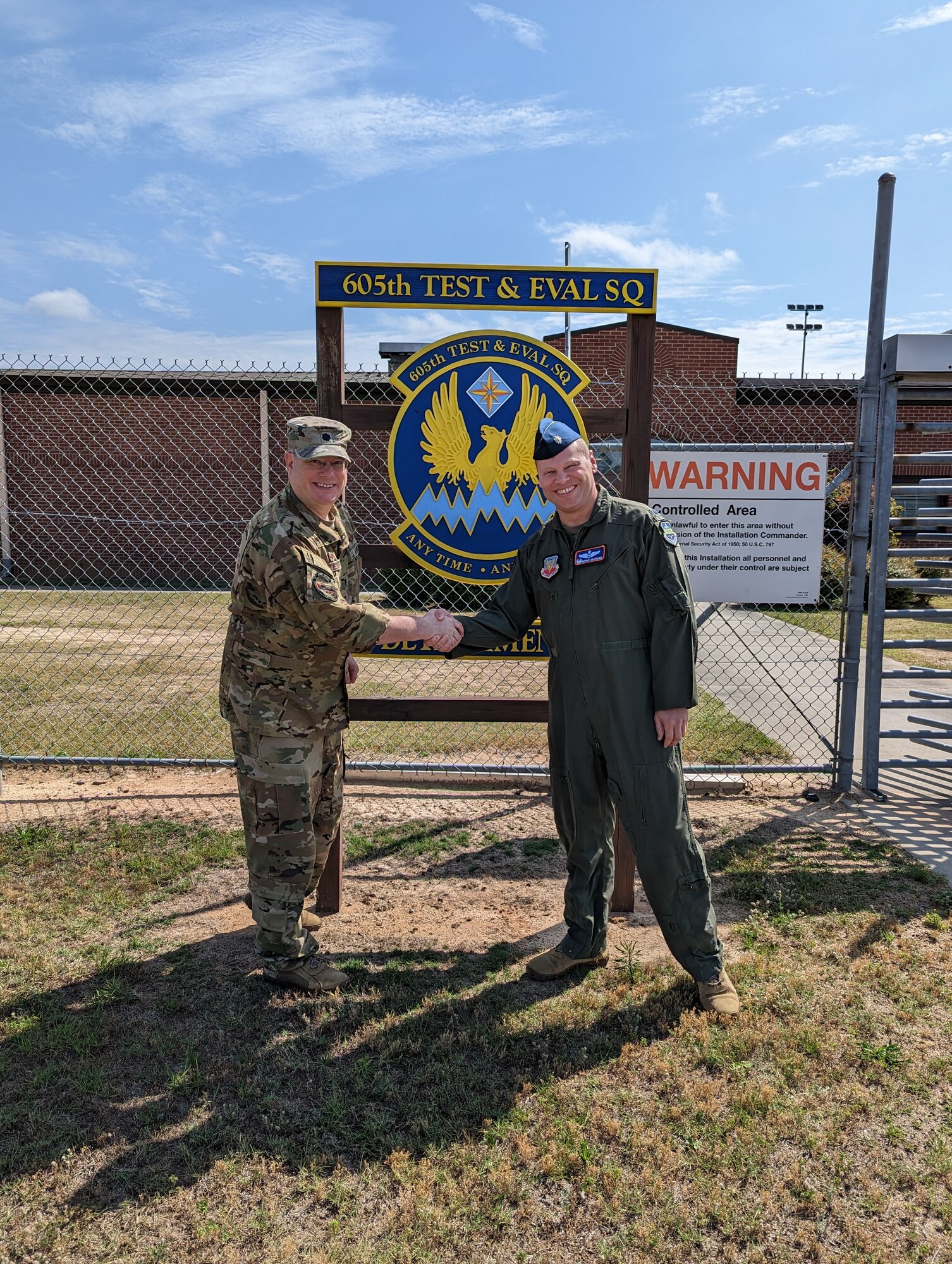photo of two uniformed U.S. Air Force Airman shaking hands outside gated area next to sign “605th Test and Evaluation Squadron Detachment 2” with unit’s emblem in the center of the sign which states “605th Test & Eval Sq, any time, any place.”
