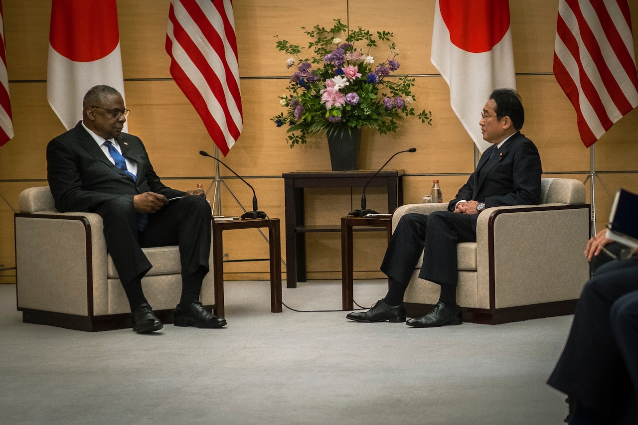 Two people face each other while seated with U.S. and Japanese flags behind them.