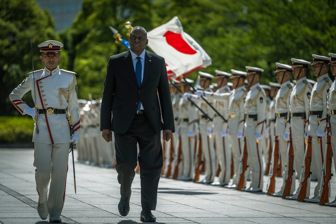 Two people walk toward the camera while Japanese service members stand in formation to the right.