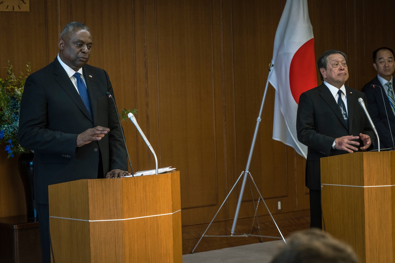 Two people in suits stand behind podiums, separated by a Japanese flag in between them.