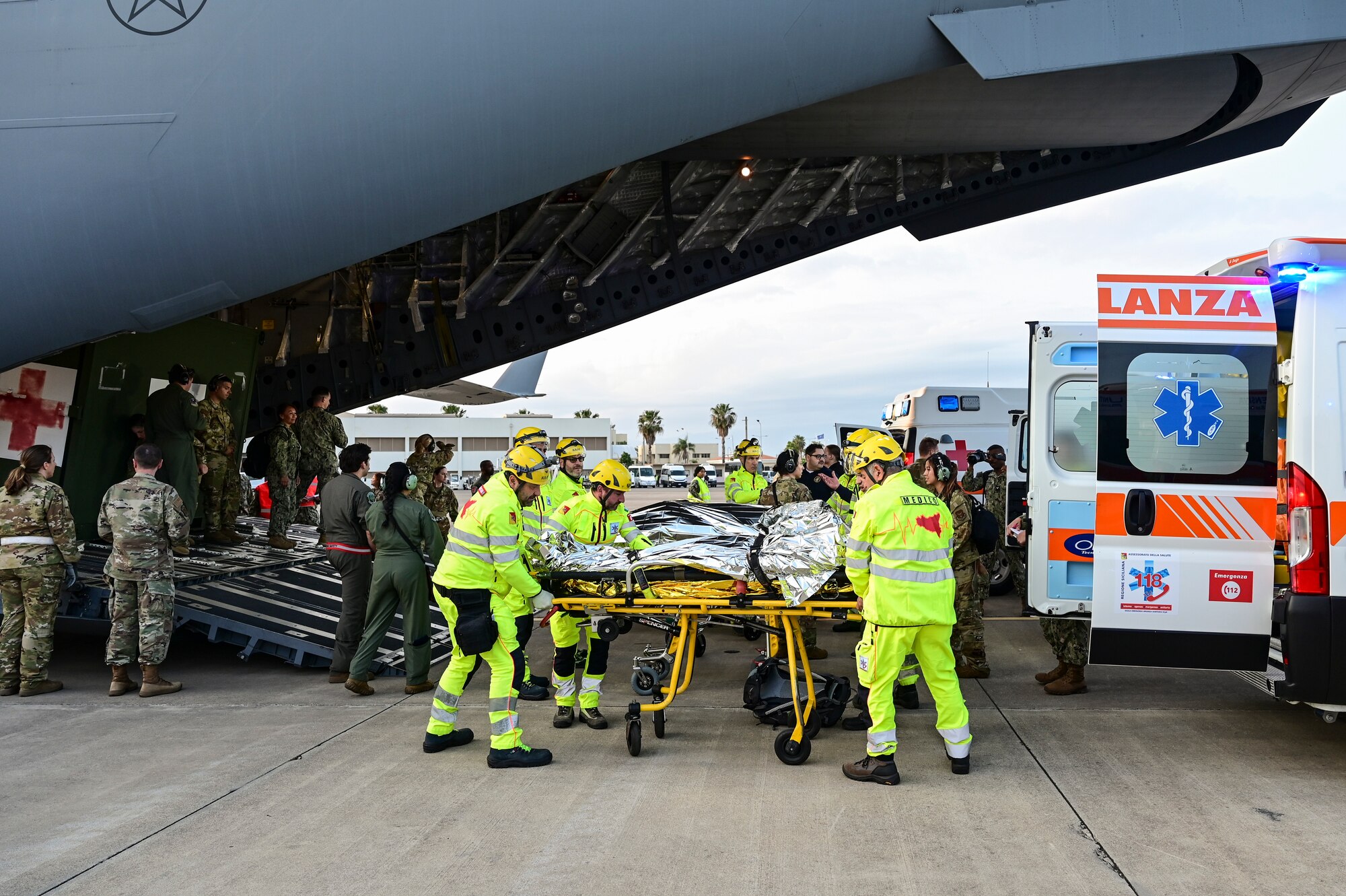 Emergency personnel in bright yellow uniforms help transfer patients from a C-17 ramp into an open ambulance on the flight line as military medical personnel look on.