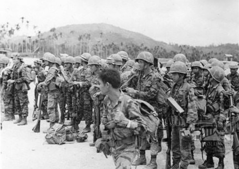 Several soldiers stand together wearing rucksacks and helmets and carrying rifles.