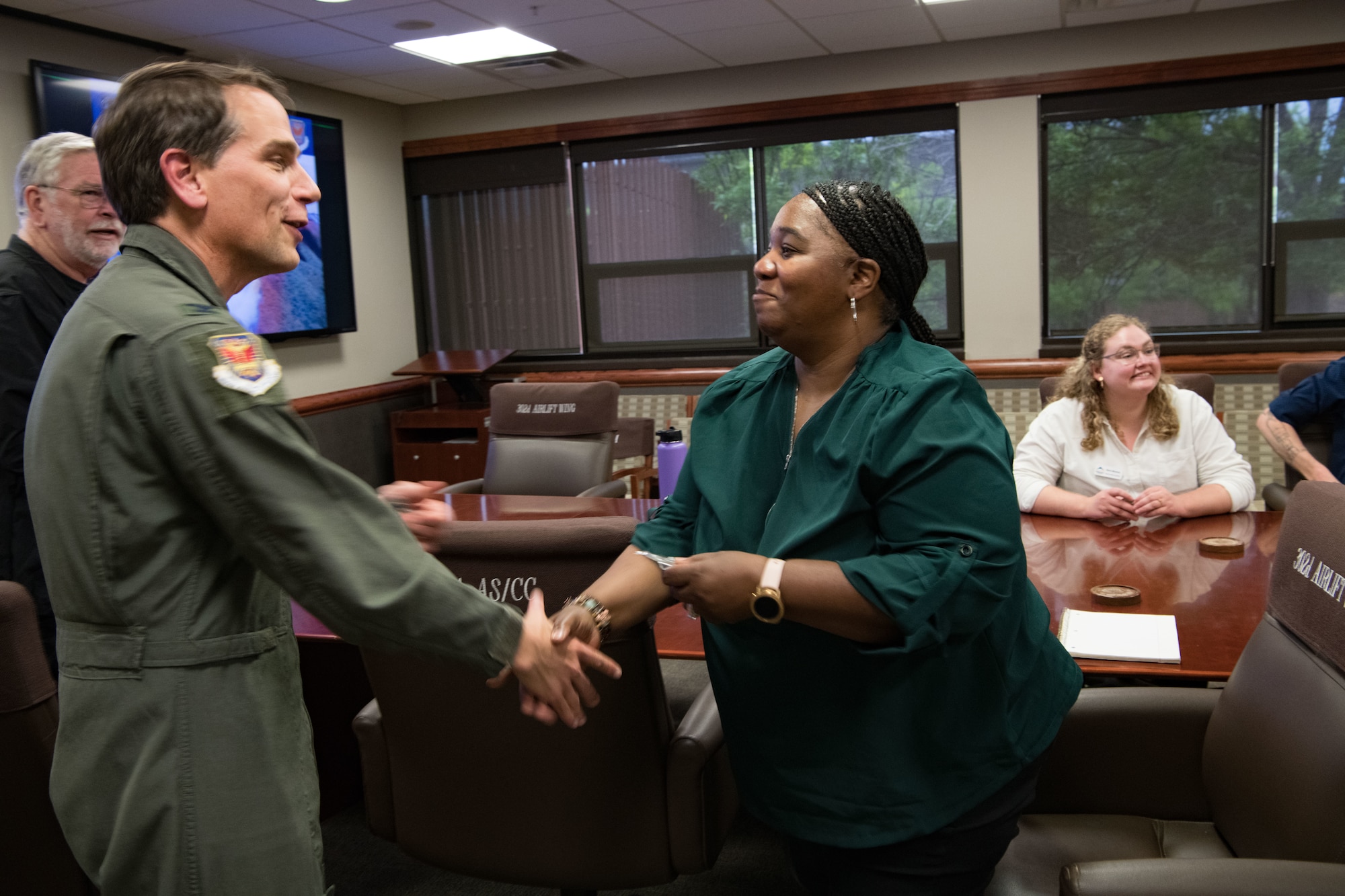 A man in a flight suit hands a civilian woman a coin and prepares to shake her hand in a conference room.