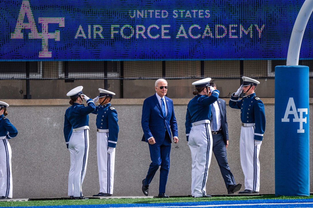 Air Force cadets salute as the president, wearing a suit and sunglasses, walks across a football field.