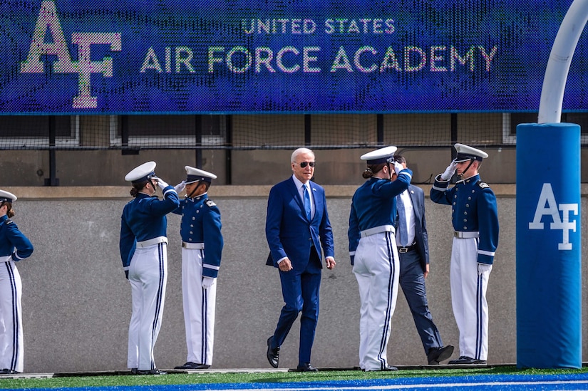 Air Force cadets salute as the president, wearing a suit and sunglasses, walks across a football field.